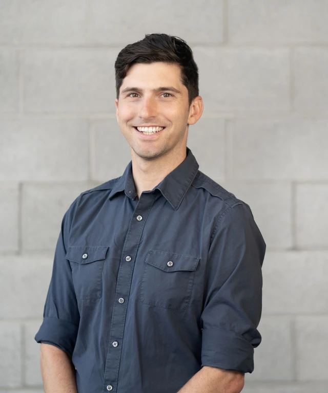 David Hallock wearing a navy button down, smiling in front of a light colored cinderblock wall.