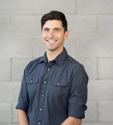 David Hallock wearing a navy button down, smiling in front of a light colored cinderblock wall.